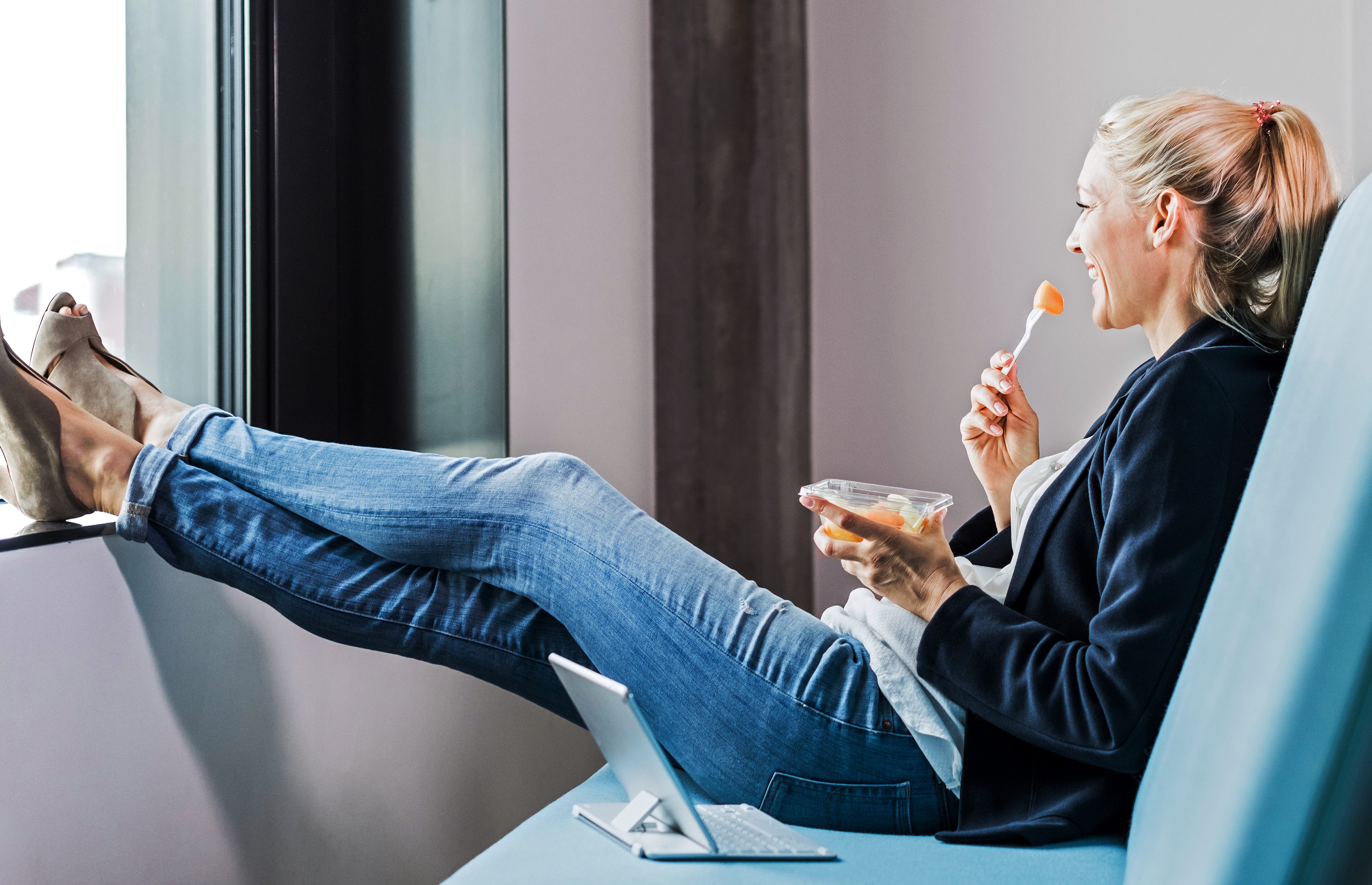 Eine Frau sitzt entspannt auf einem Sofa. Sie hält eine Schale mit Essen in der Hand und isst mit einer Gabel. Vor ihr steht ein aufgeklappter Laptop.