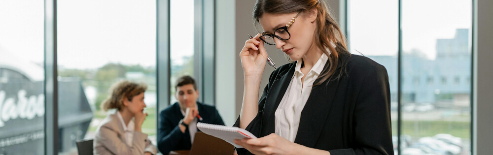 Eine Geschäftsfrau in einem dunklen Anzug und mit Brille steht in einem modernen Bürogebäude und sieht auf ein Notizbuch in ihrer Hand. Im Hintergrund sind zwei Personen in Businesskleidung zu sehen, die an einem Tisch sitzen und miteinander sprechen. 