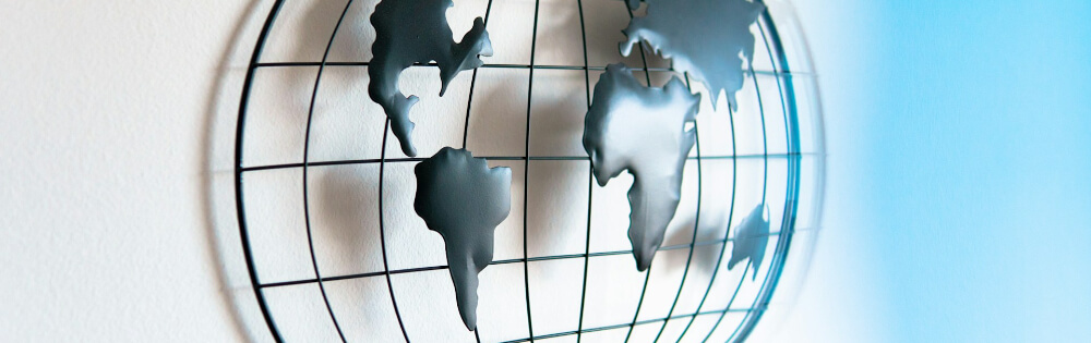 Auslandskranken-Gruppenversicherung Ein kunstvolles Metallgitter in Form einer Weltkarte hängt an einer weißen Wand. Die Kontinente sind als flache, dunkle Silhouetten dargestellt, die an einem runden Gitterrahmen befestigt sind.